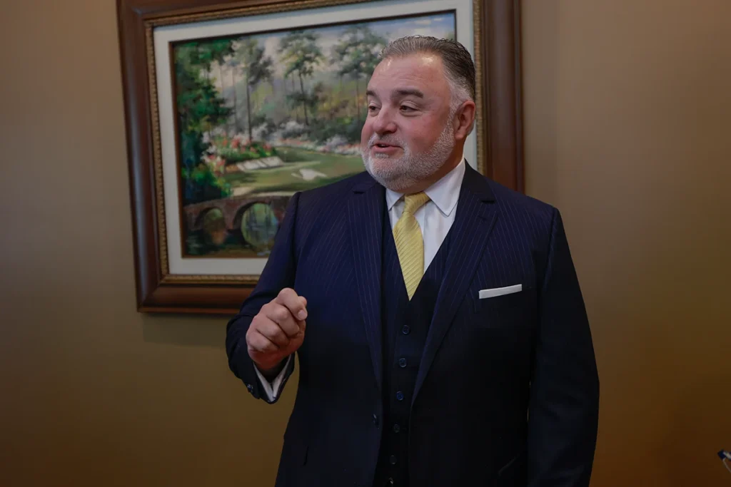Lawyer Tom McCutcheon gestures while speaking about financial recovery for motor vehicle crash victims. A painting is in the.