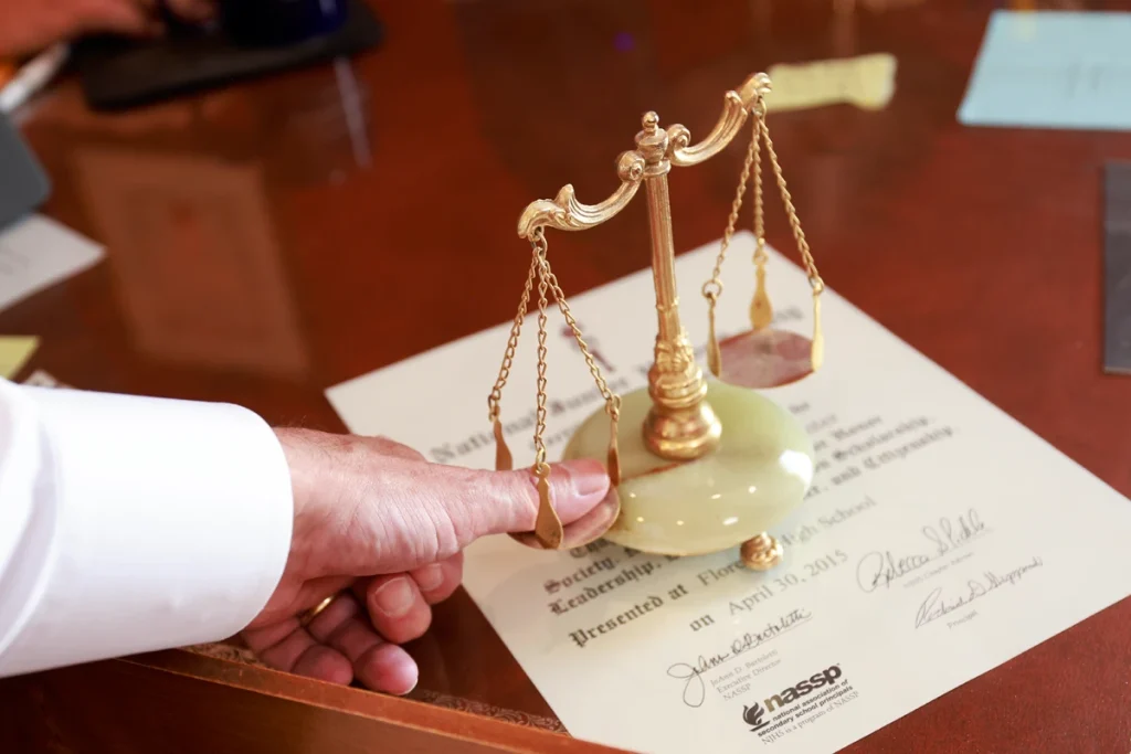 A hand touches a small gold scale of justice on a desk with a certificate and papers in the background.